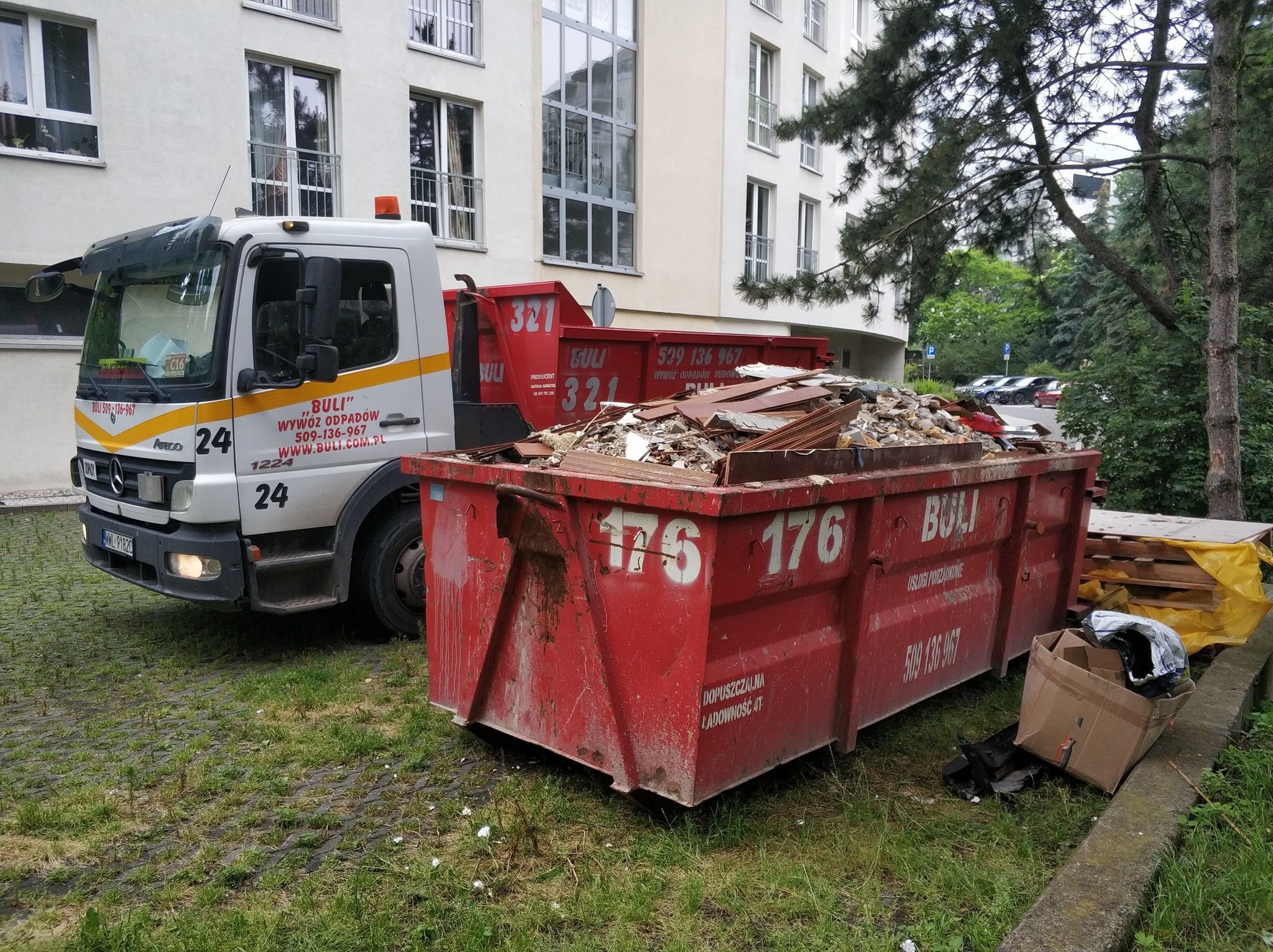 Czerwony kontener wypełniony gruzem budowlanym stoi obok białej ciężarówki z napisem 'Buli - Wywóz odpadów' na tle budynku mieszkalnego i zieleni.