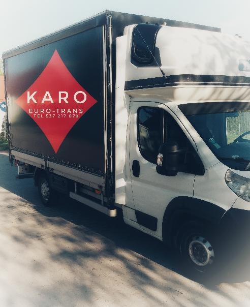 Biały bus dostawczy z czarną plandeką i dużym czerwonym logo w kształcie rombu z napisem KARO EURO-TRANS i numerem telefonu, zaparkowany na zewnątrz.