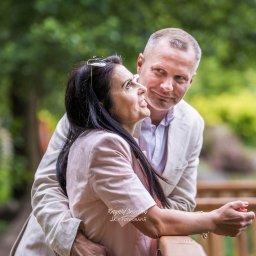 JK-Fotografia Krzysztof Jastrzębski - Zakochana para w jasnych strojach, oparta o drewnianą balustradę, spogląda na siebie z czułością na tle zieleni. Romantyczna sesja plenerowa.