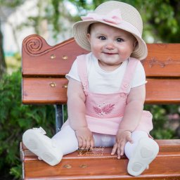 JK-Fotografia Krzysztof Jastrzębski - Uśmiechnięta dziewczynka w kapeluszu z kokardą siedzi na drewnianej ławce w parku, ubrana w różową sukienkę i białe buciki. Widoczne logo fotografa.