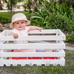 JK-Fotografia Krzysztof Jastrzębski - Uśmiechnięta dziewczynka w słomkowym kapeluszu siedzi w białej, drewnianej skrzynce na tle zielonego ogrodu. Urocza sesja plenerowa z dzieckiem.