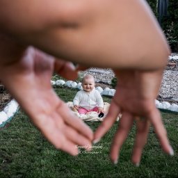 JK-Fotografia Krzysztof Jastrzębski - Uśmiechnięte niemowlę siedzi na kocu w ogrodzie, sfotografowane przez dłonie tworzące ramę w kształcie serca. Jasne, naturalne światło.