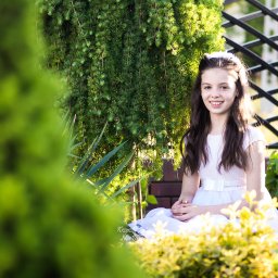 JK-Fotografia Krzysztof Jastrzębski - Uśmiechnięta dziewczynka w białej sukience, siedząca w ogrodzie na tle zieleni i metalowej konstrukcji. Delikatne światło podkreśla jej twarz.