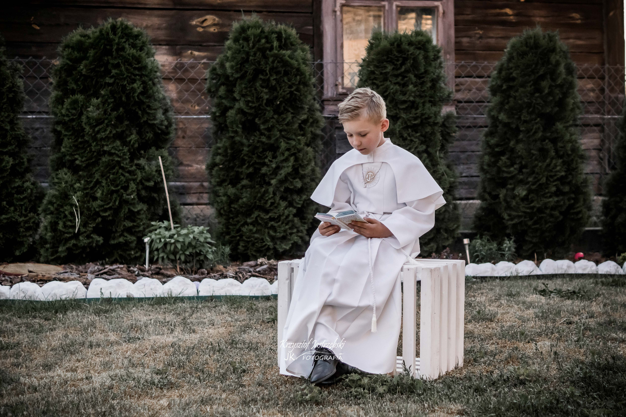Chłopiec w stroju komunijnym siedzi na białej skrzynce, czytając książeczkę. W tle drewniany dom i tuje. Ujęcie z dołu, podkreślające uroczysty charakter.