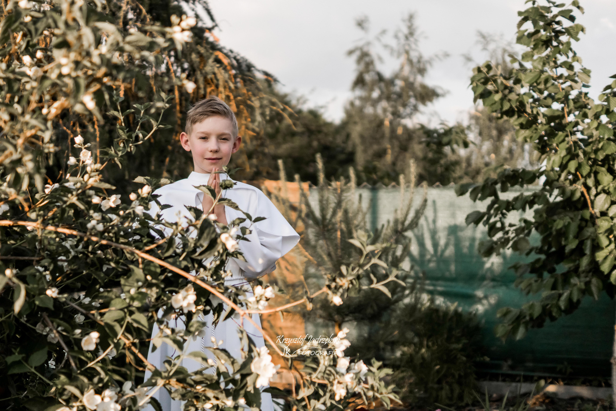 Chłopiec w stroju komunijnym, z rękami złożonymi do modlitwy, pozuje w ogrodzie pełnym kwitnących krzewów. Delikatne światło podkreśla uroczysty charakter chwili.
