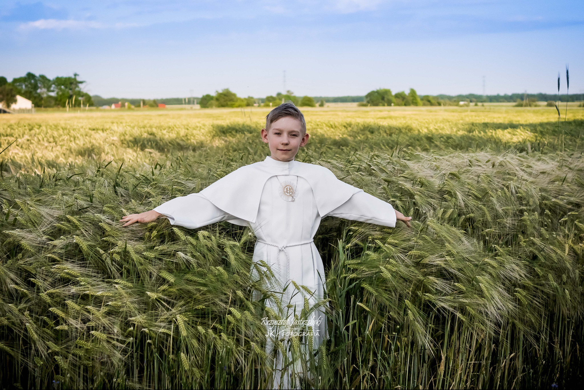 Chłopiec w stroju komunijnym stoi na polu zboża z rozłożonymi ramionami, na tle zieleni i błękitnego nieba. Ujęcie z bliska, naturalne światło.
