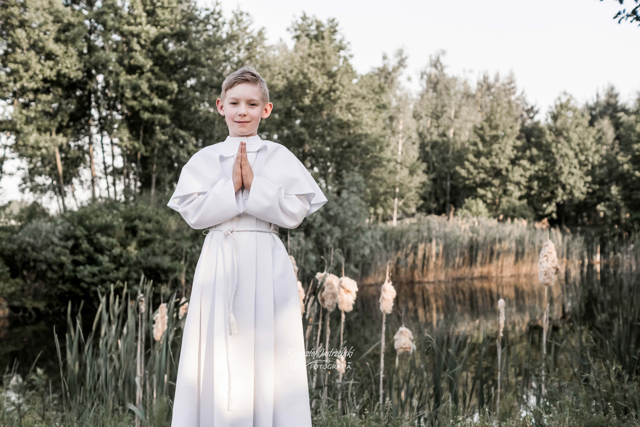 Chłopiec w albie komunijnej, z rękami złożonymi do modlitwy, na tle jeziora i trzcin, uchwycony w naturalnym świetle podczas sesji zdjęciowej.
