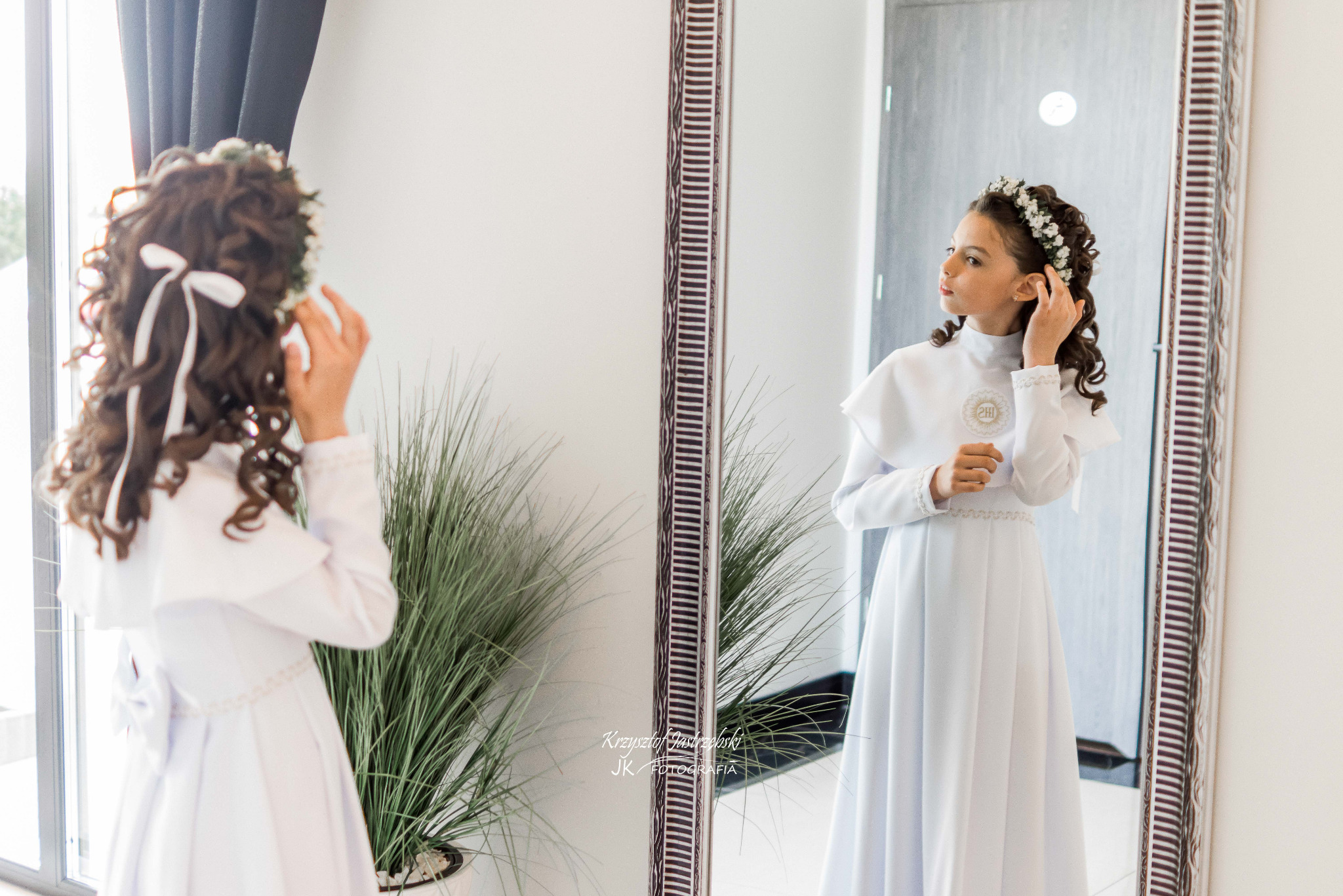 Dziewczynka w białej sukience komunijnej, z wiankiem na głowie, przegląda się w lustrze w jasnym wnętrzu. Ujęcie zza pleców, widoczny fragment rośliny doniczkowej.