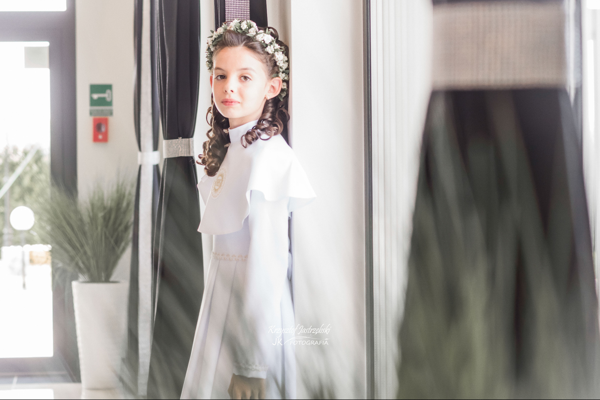 Dziewczynka w białej albie z wiankiem na głowie, pozuje w jasnym wnętrzu, obok zasłon z połyskującym pasem. Widoczny fragment rośliny doniczkowej.