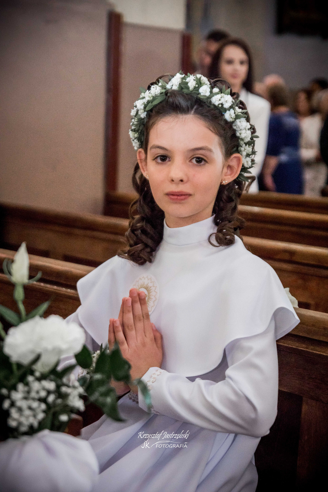 Dziewczynka w białej albie i wianku, z złożonymi dłońmi, siedzi w ławce kościoła podczas uroczystości Pierwszej Komunii Świętej. W tle wierni.