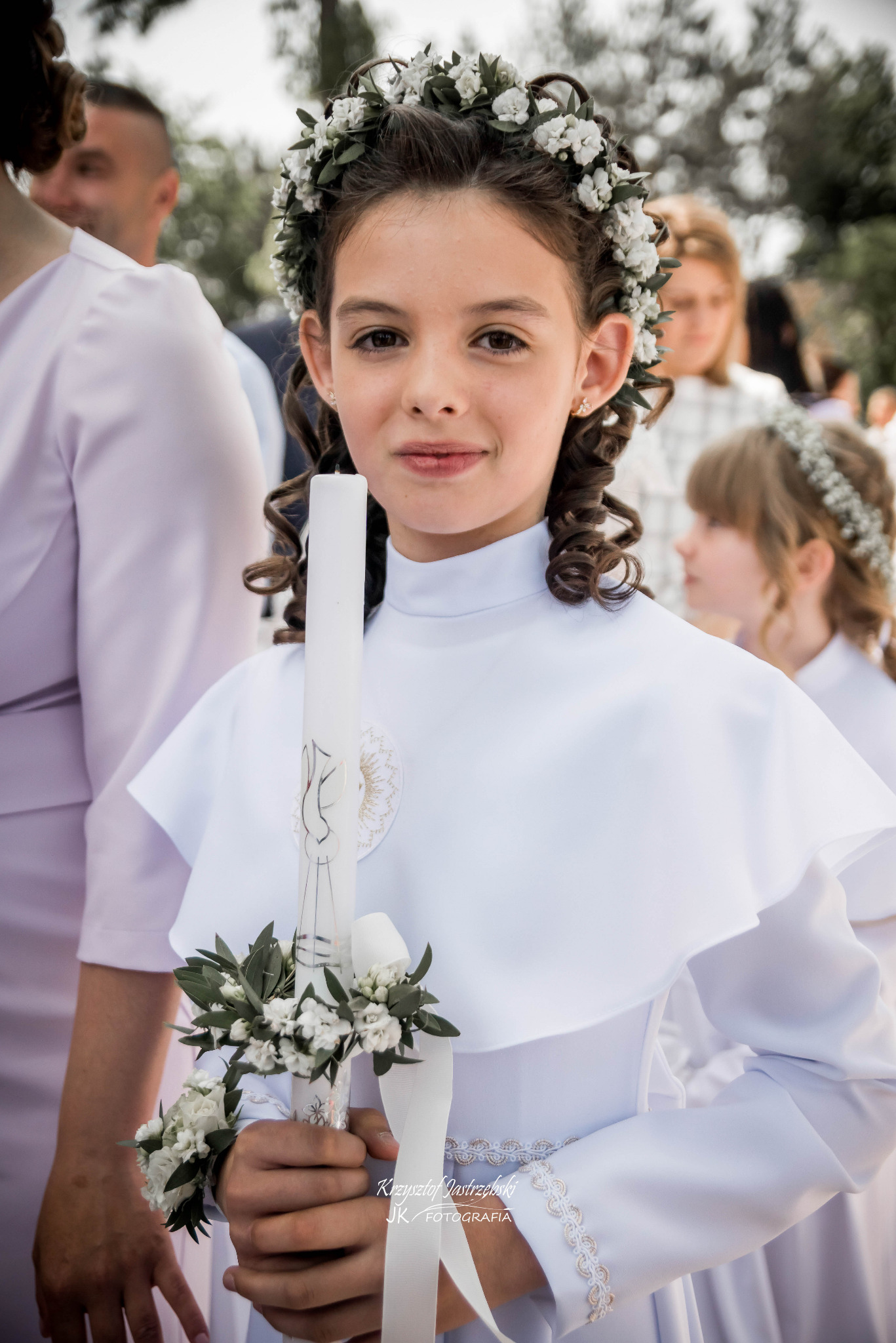 Portret dziewczynki w białej sukience komunijnej z wiankiem i świecą, na tle innych uczestników uroczystości. Delikatne oświetlenie, skupienie na twarzy dziecka.