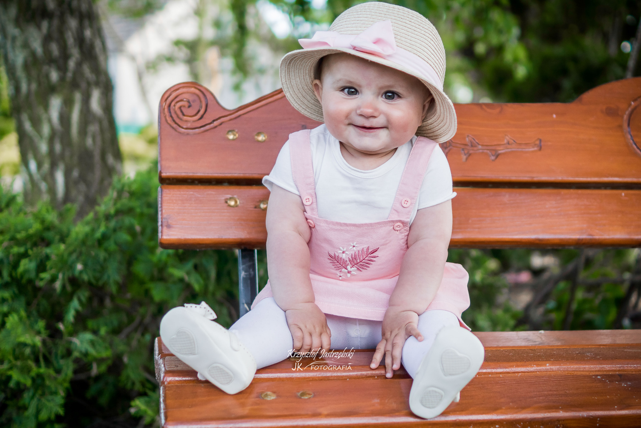 Uśmiechnięta dziewczynka w kapeluszu z kokardą siedzi na drewnianej ławce w parku, ubrana w różową sukienkę i białe buciki. Widoczne logo fotografa.
