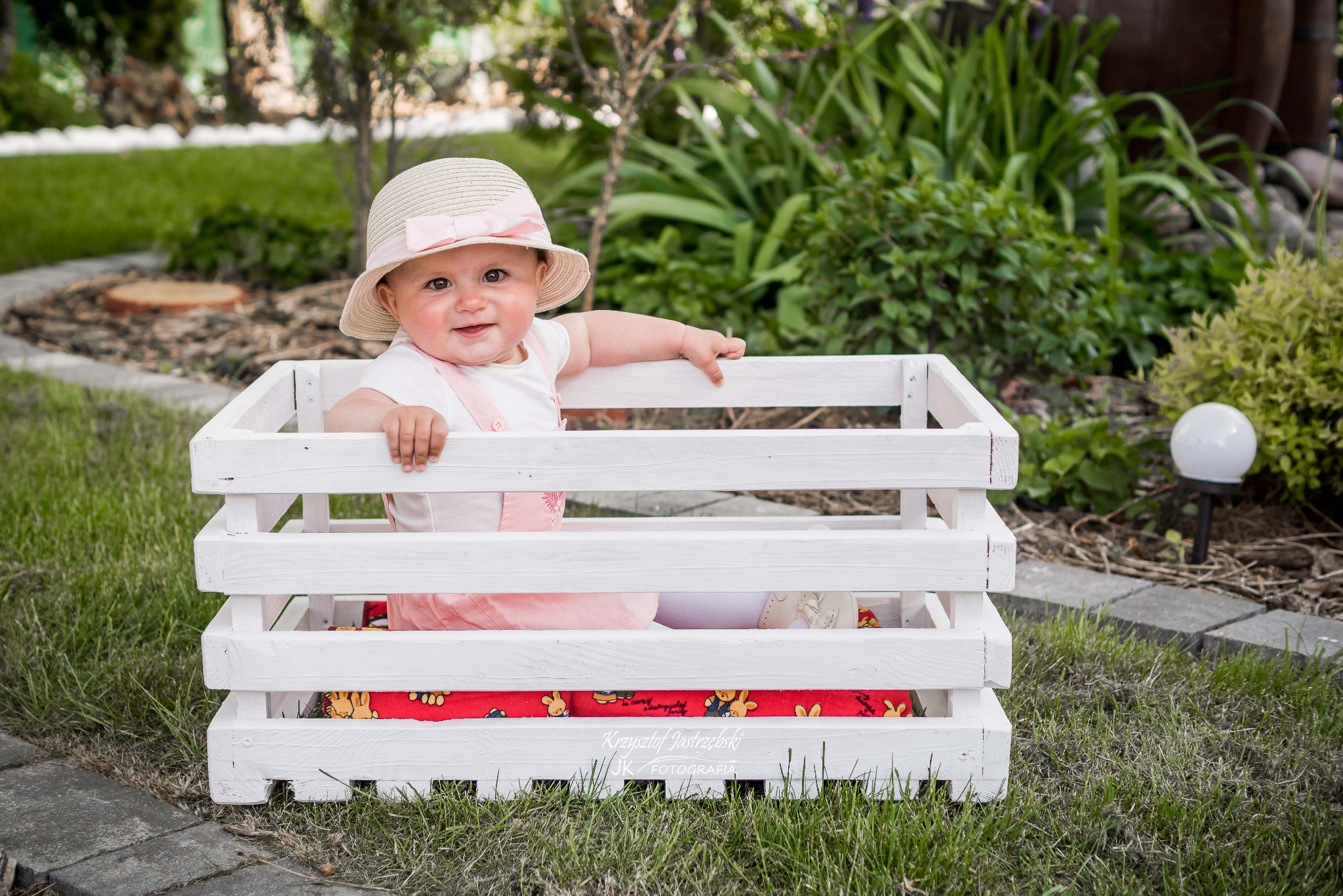Uśmiechnięta dziewczynka w słomkowym kapeluszu siedzi w białej, drewnianej skrzynce na tle zielonego ogrodu. Urocza sesja plenerowa z dzieckiem.
