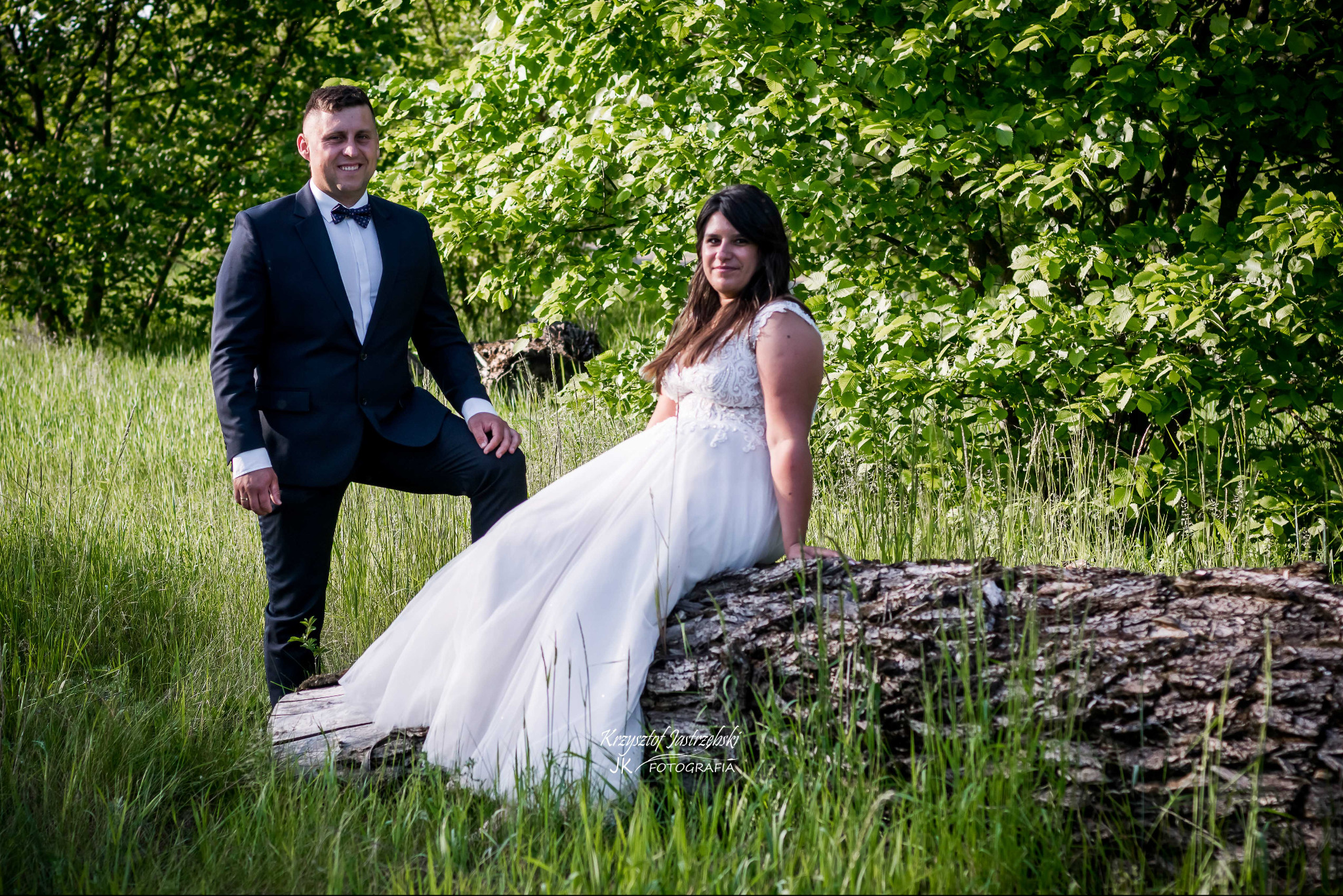 Para młoda w plenerze: Pan młody w garniturze opiera się o kłodę, Panna młoda w sukni siedzi na niej, za nimi drzewa i trawa. Romantyczna sesja w naturalnym otoczeniu.