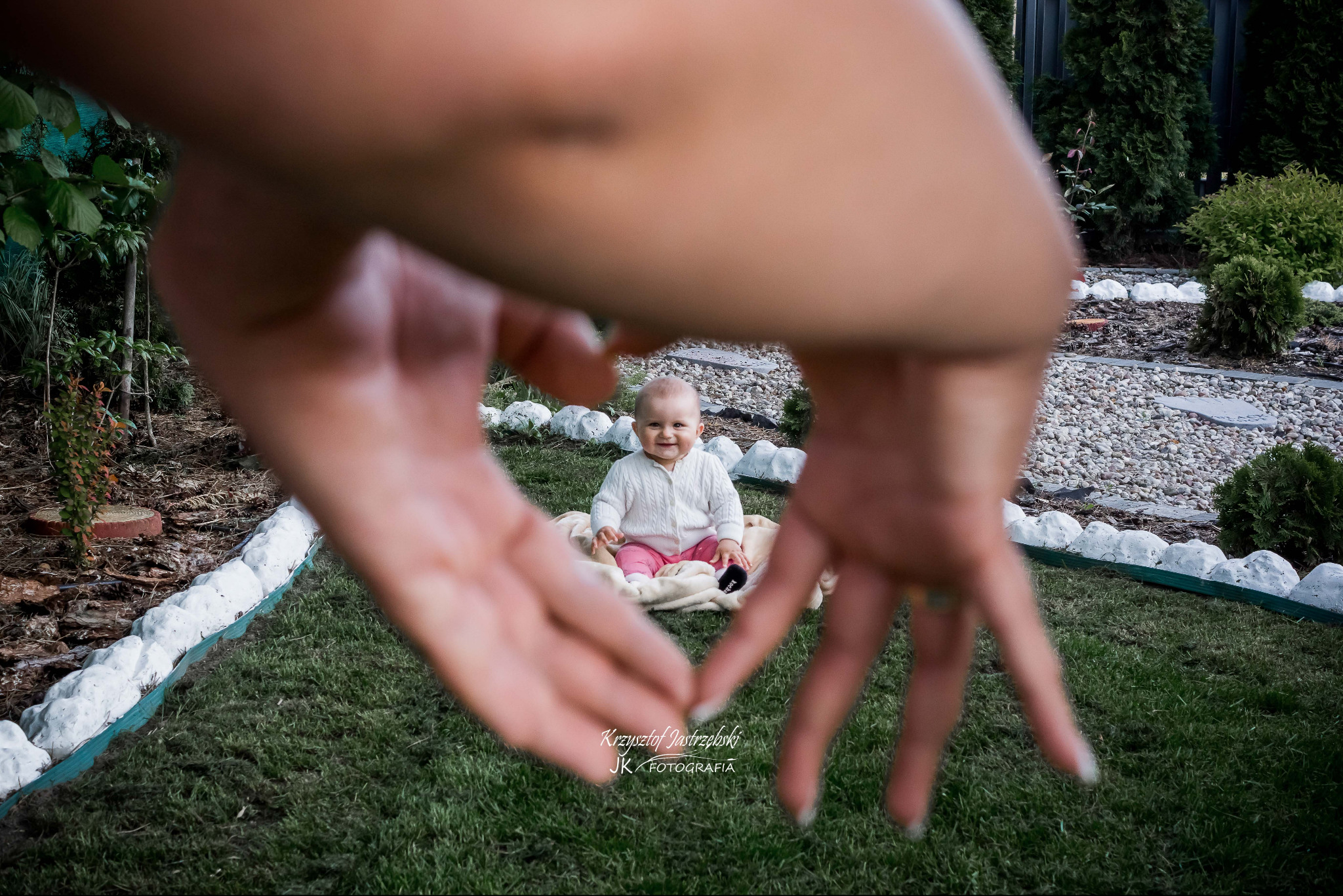 Uśmiechnięte niemowlę siedzi na kocu w ogrodzie, sfotografowane przez dłonie tworzące ramę w kształcie serca. Jasne, naturalne światło.