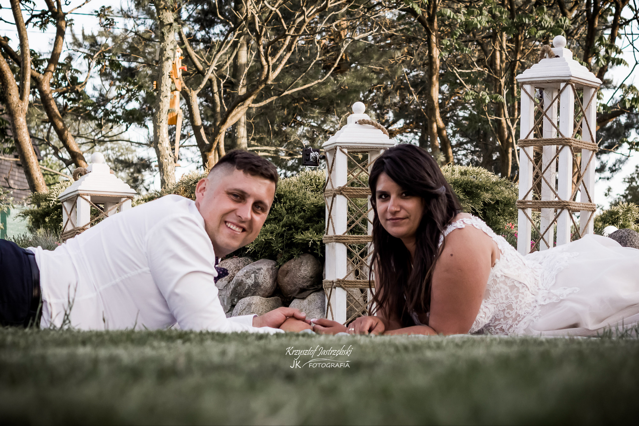 Uśmiechnięta para młoda leży na trawie, trzymając się za ręce. W tle dekoracje ślubne i drzewa, tworząc romantyczną scenerię.