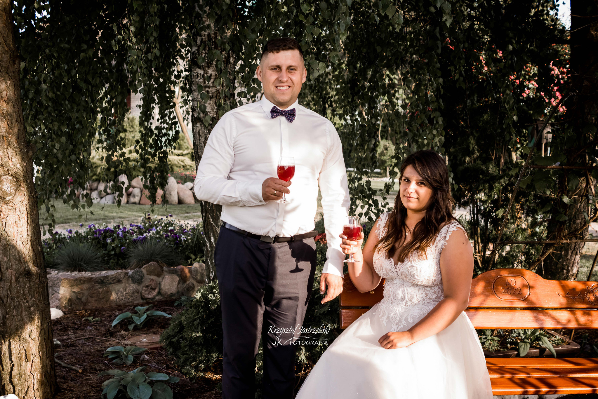 Para młoda pozuje z kieliszkami czerwonego wina na tle zieleni. Panna młoda w białej sukni siedzi na ławce, pan młody stoi obok w białej koszuli i muszce.