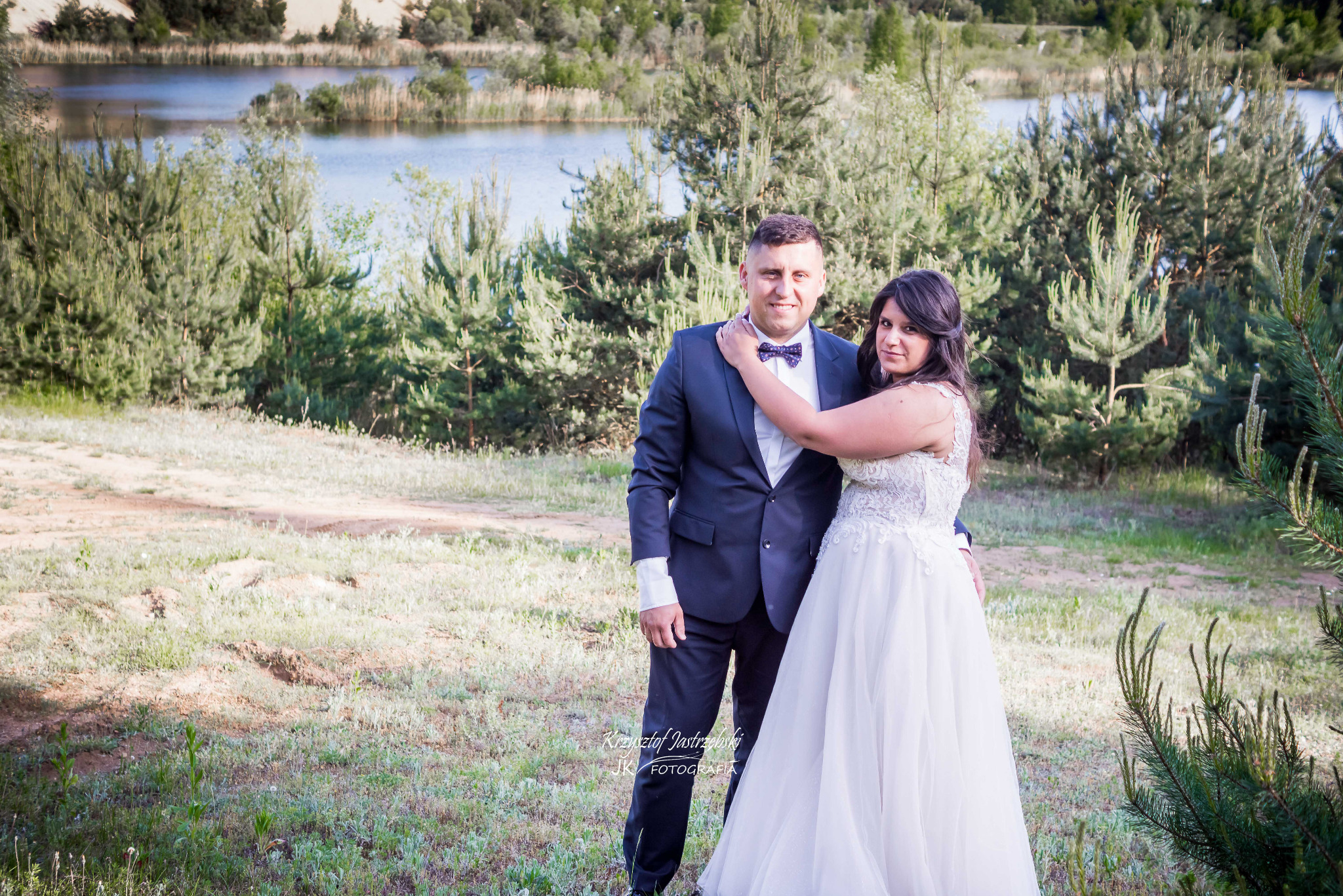 Para młoda w plenerze: pan młody w granatowym garniturze, panna młoda w białej sukni, jezioro i las w tle, naturalne światło, ujęcie w zieleni.