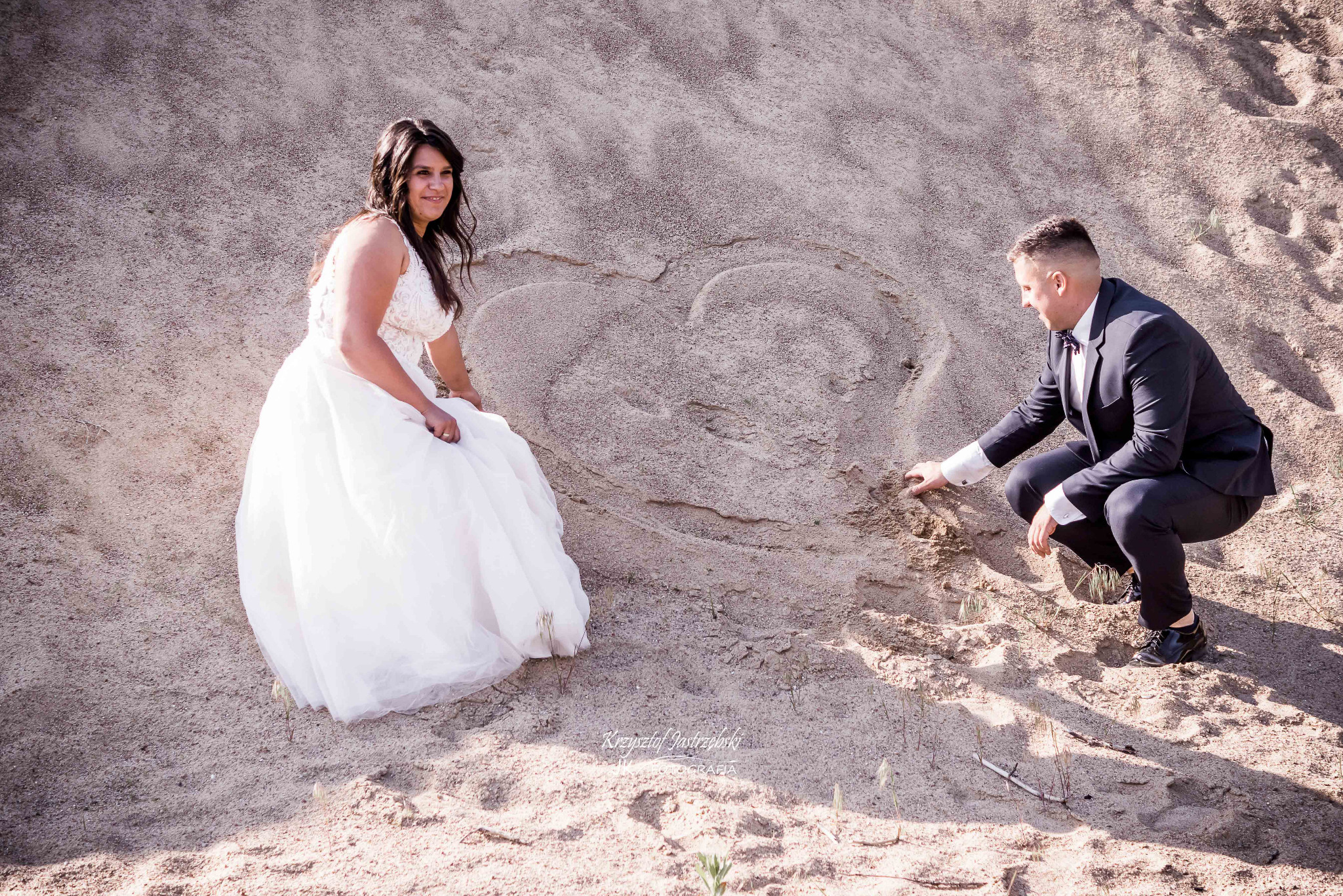 Para młoda rysuje serce na piasku. Panna młoda w białej sukni spogląda w bok, pan młody w garniturze klęczy i rysuje palcem serce. Naturalne światło, piaszczyste tło.