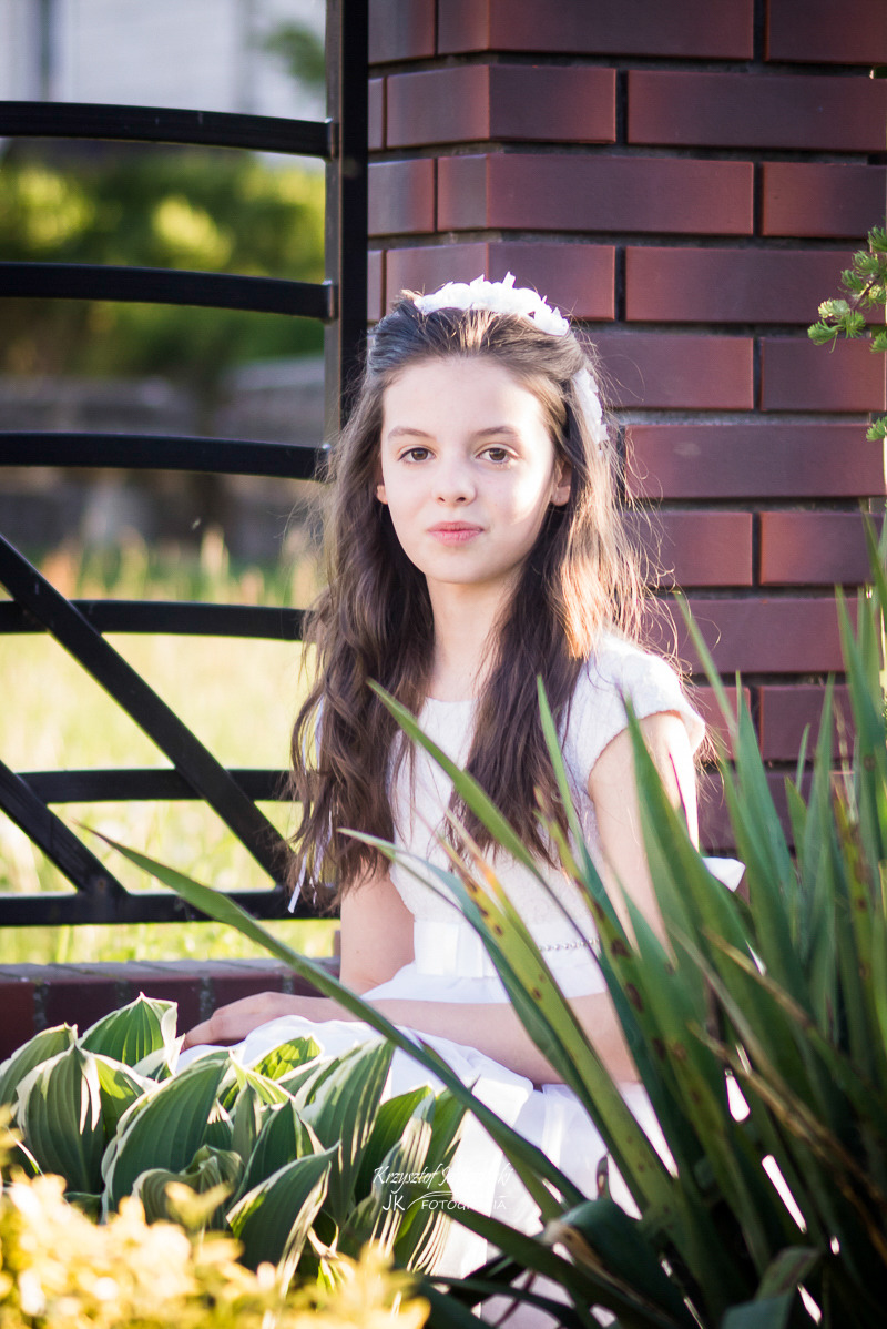 Dziewczynka w białej sukience i wianku siedzi przy ceglanym murku, spoglądając w obiektyw. Ujęcie z dołu, z roślinami w pierwszym planie. Delikatne światło.