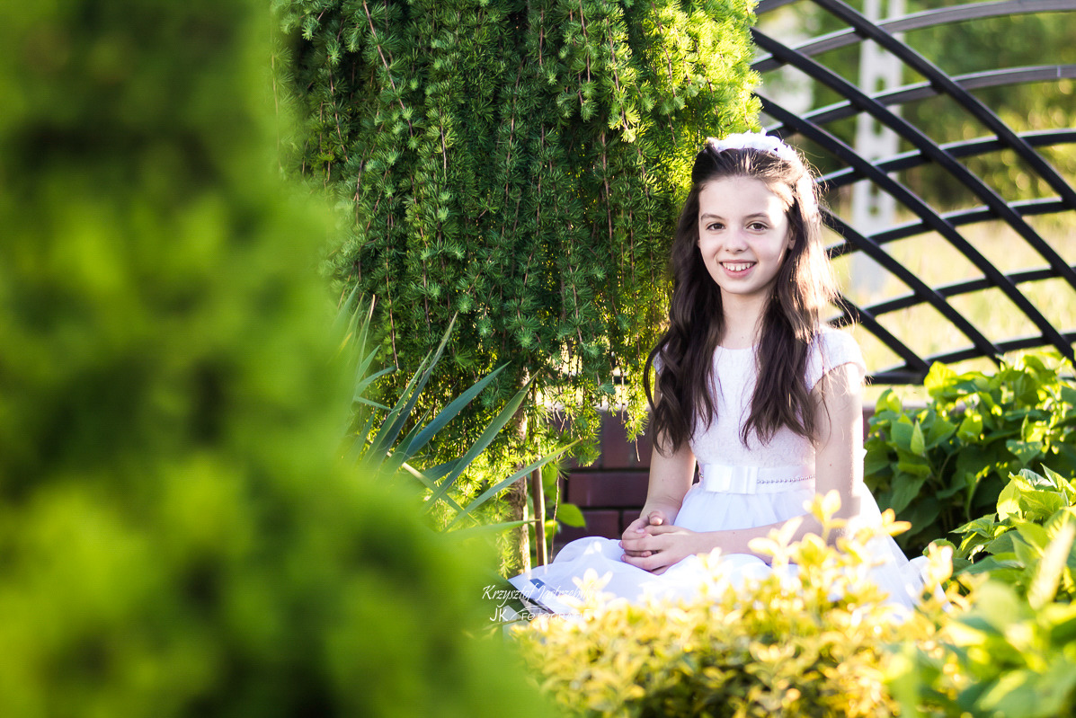 Uśmiechnięta dziewczynka w białej sukience, siedząca w ogrodzie na tle zieleni i metalowej konstrukcji. Delikatne światło podkreśla jej twarz.
