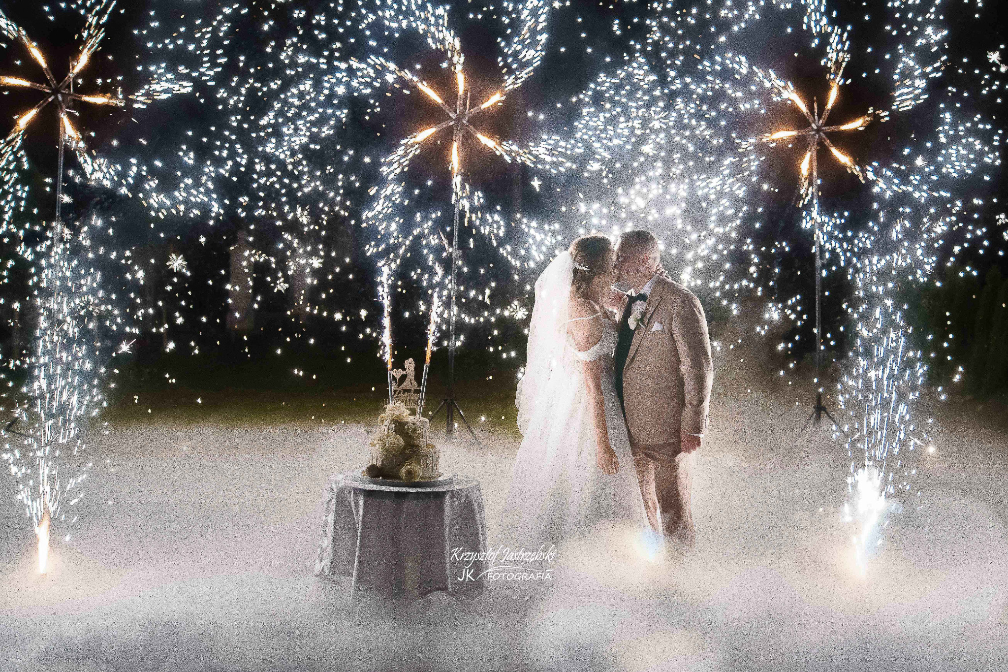 Pocałunek pary młodej na tle nocnego nieba rozświetlonego sztucznymi ogniami i tortu weselnego na stole. Romantyczna atmosfera.