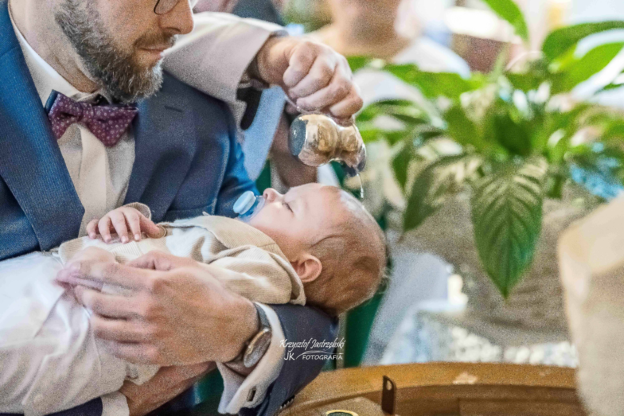 Ujęcie z ceremonii chrztu: mężczyzna w garniturze trzyma śpiące niemowlę z smoczkiem, nad którym kapłan polewa wodą z naczynia. W tle rośliny doniczkowe.