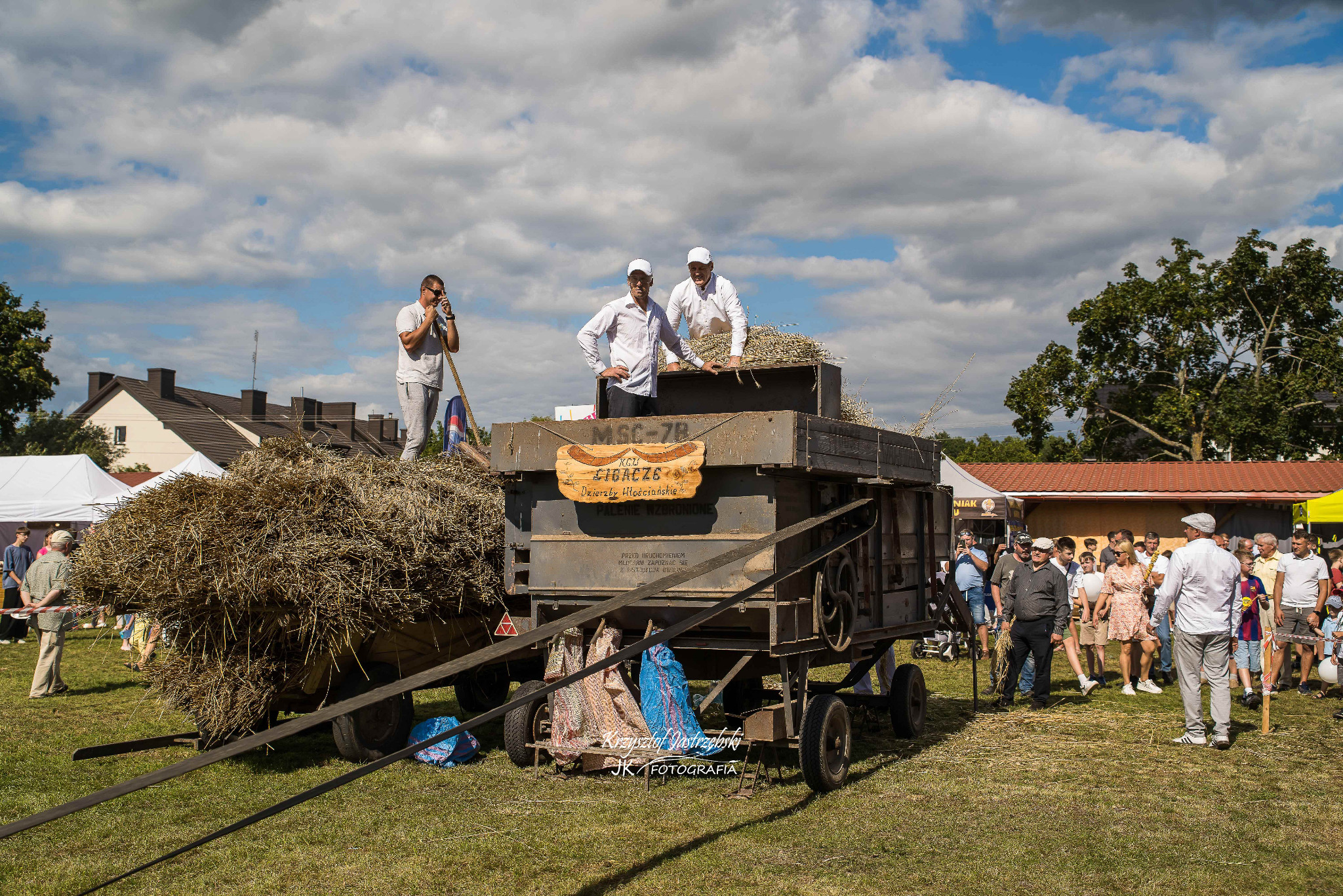 Plenerowe widowisko z młockarnią MSC-7B, mężczyźni w białych koszulach prezentują pracę maszyny podczas festynu. Tłum obserwatorów w tle, słoneczna pogoda.