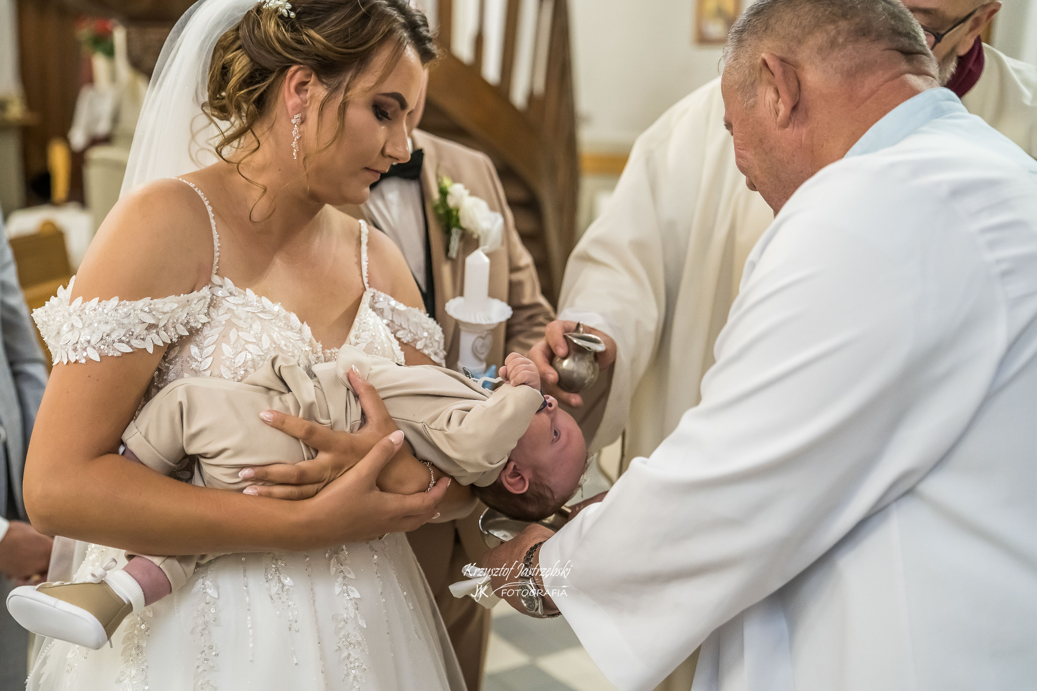 Uroczystość chrztu: młoda mama w sukni ślubnej trzyma dziecko podczas ceremonii. Ksiądz polewa główkę dziecka wodą. W tle goście w eleganckich strojach.