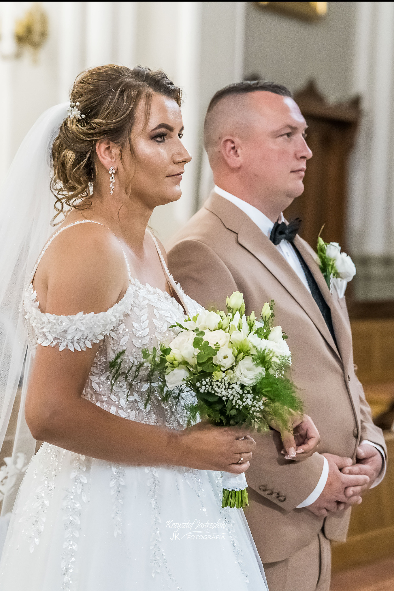 Panna młoda w zdobionej sukni ślubnej trzyma bukiet, obok pan młody w beżowym garniturze z muchą, oboje patrzą w prawo. Para podczas ceremonii ślubnej.
