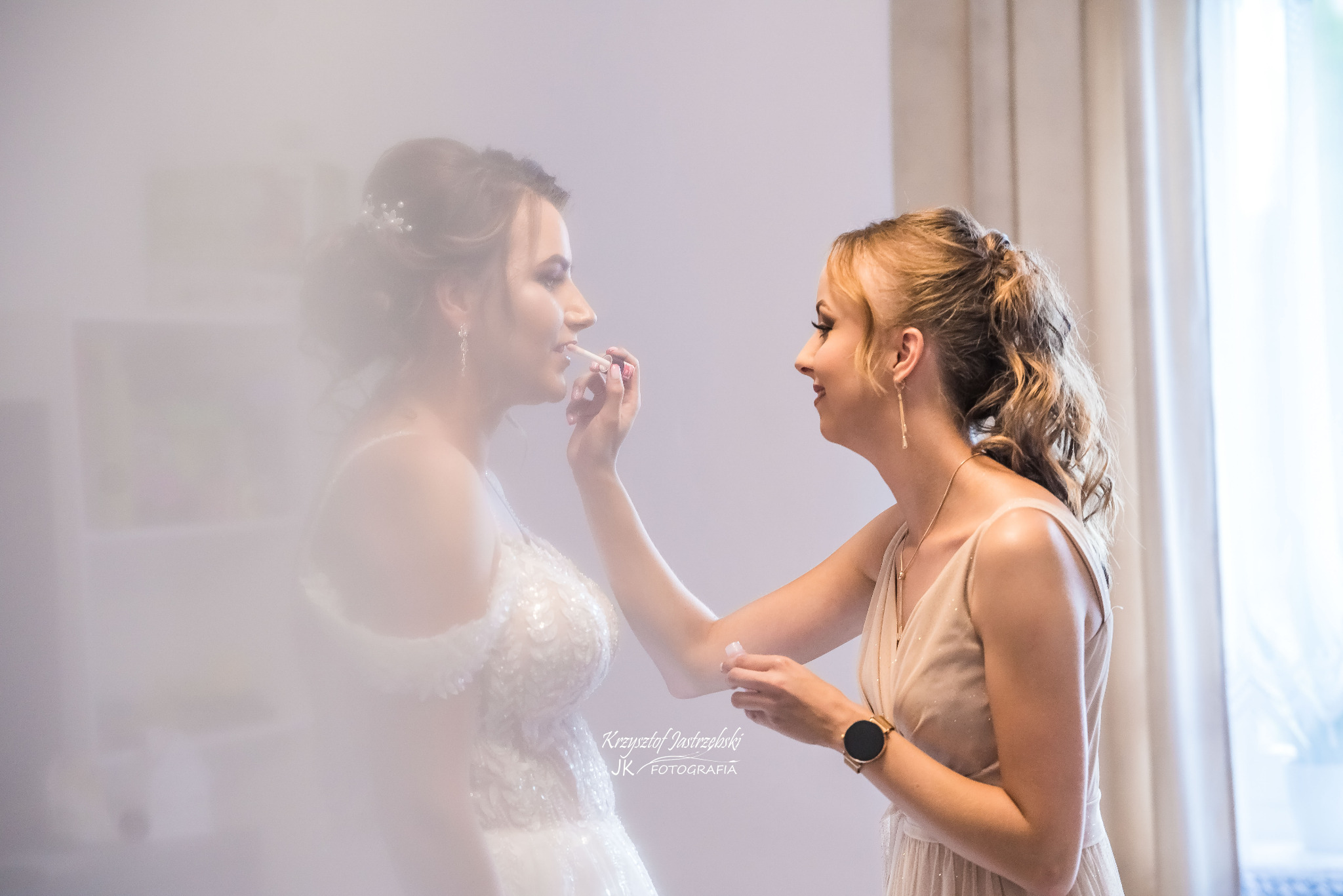 Panna młoda w sukni ślubnej malowana szminką przez kobietę w sukience, jasne oświetlenie, delikatny efekt rozmycia, przygotowania do ślubu.