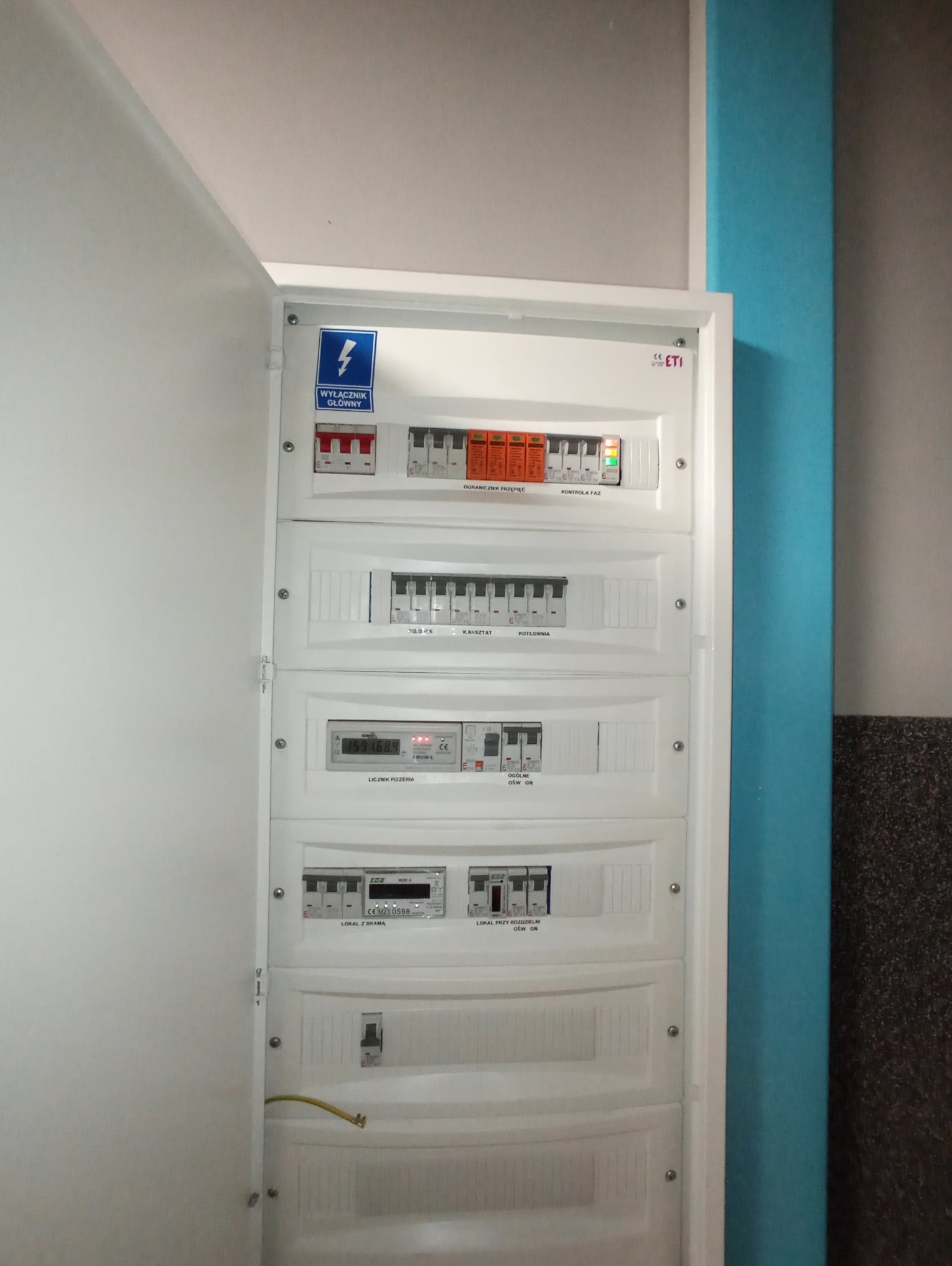 Otwarta skrzynka rozdzielcza z widocznymi zabezpieczeniami przepięciowymi, wyłącznikami, licznikiem energii elektrycznej i oznaczeniami obwodów, zamontowana na ścianie z niebieskim pionowym pasem.