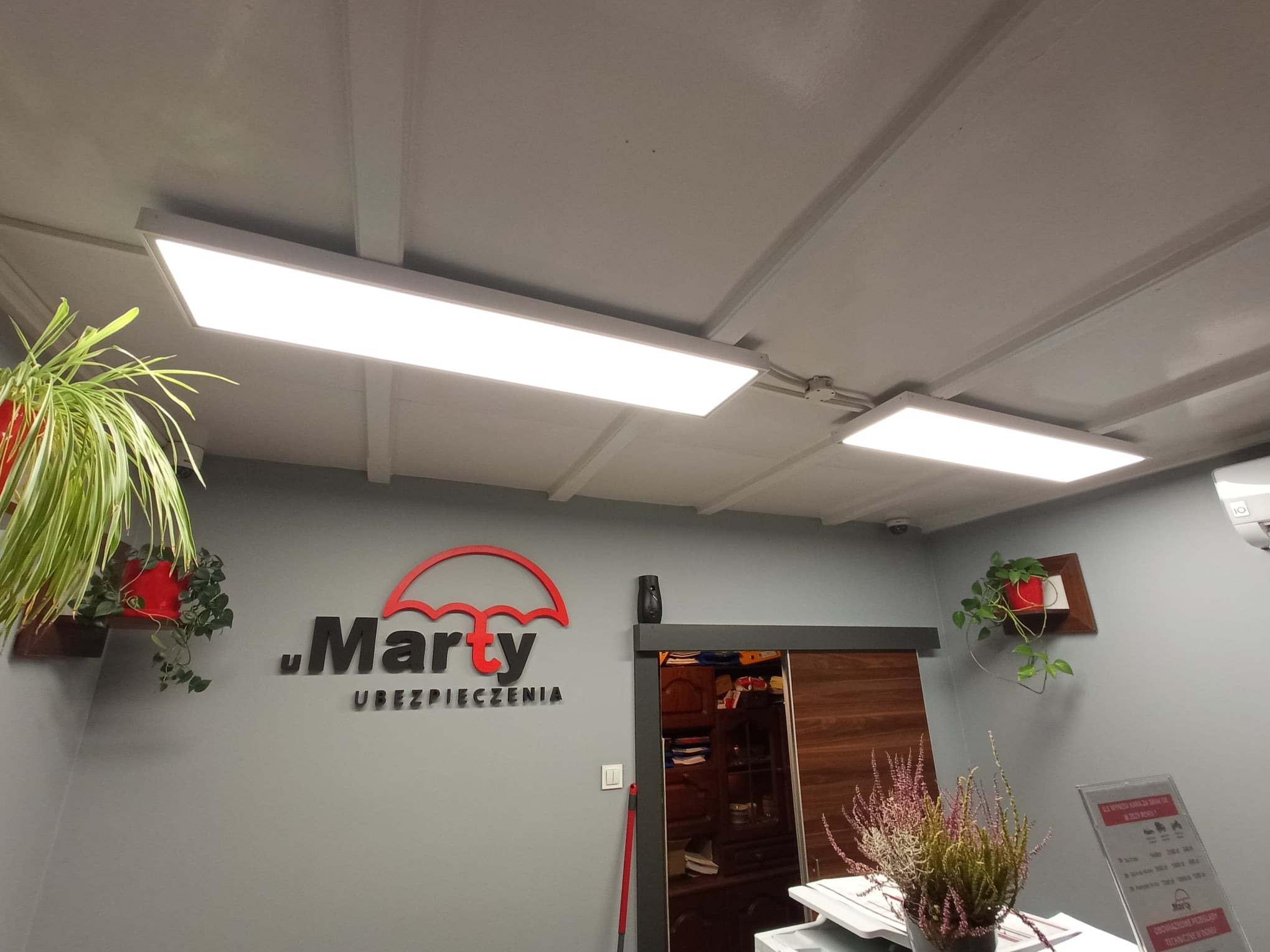 Biuro ubezpieczeń z dwoma podwieszanymi panelami LED oświetlającymi sufit, widoczne rośliny doniczkowe na ścianach i logo firmy 'Marty Ubezpieczenia'.