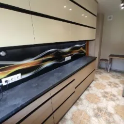 Nowoczesna zabudowa kuchenna z beżowymi szafkami górnymi z czarnymi obramowaniami i drewnopodobnymi szafkami dolnymi z szarym blatem oraz dekoracyjnym panelem szklanym w kuchni w Las.