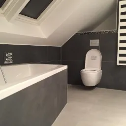 Nowoczesna łazienka na poddaszu z wanną, podwieszaną toaletą i grzejnikiem drabinkowym na tle szarych płytek, doświetlona oknem dachowym.