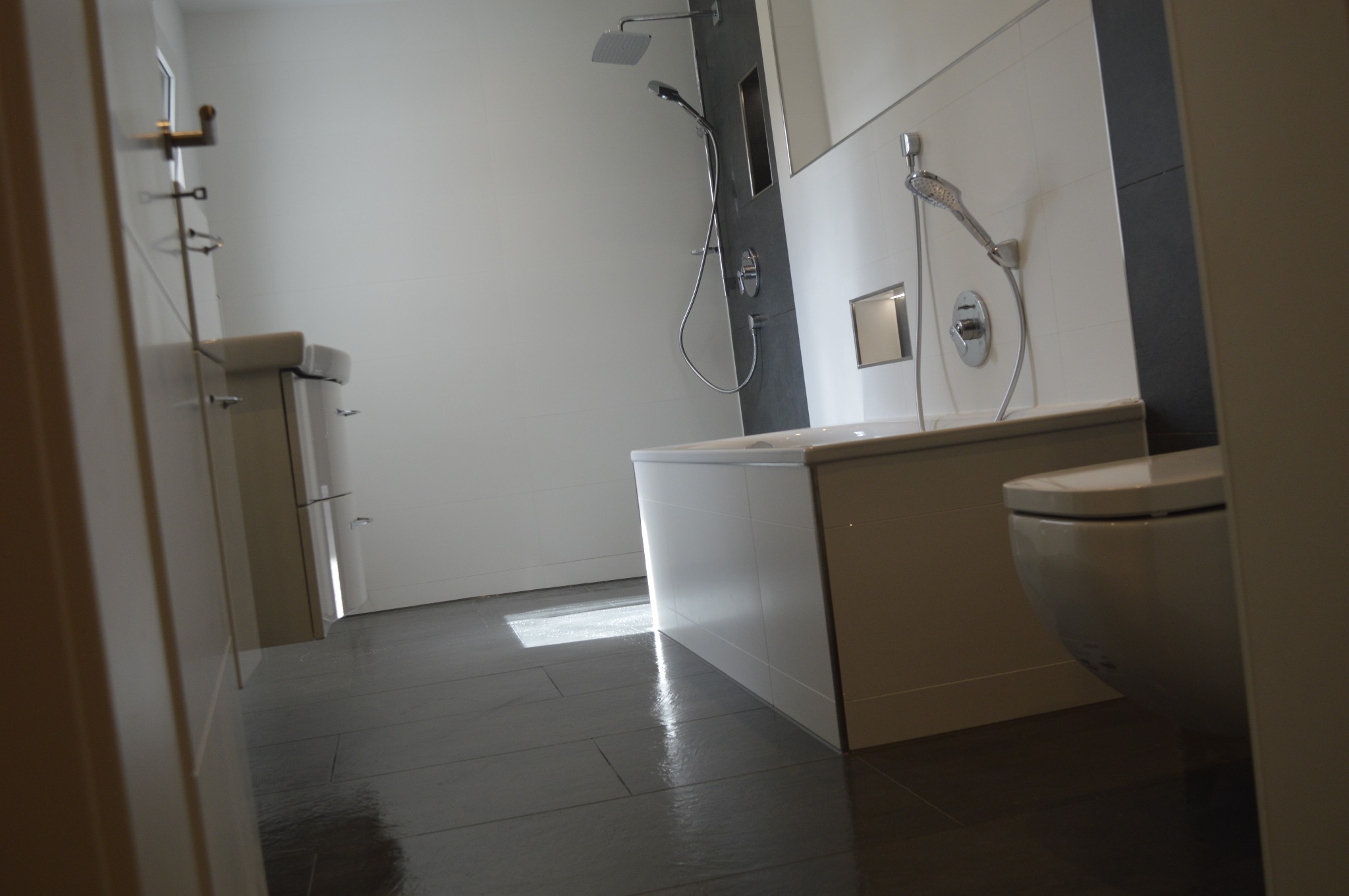 Nowoczesna łazienka z szarymi płytkami podłogowymi, białą wanną obudowaną płytkami, wiszącą toaletą i kabiną prysznicową z deszczownicą.