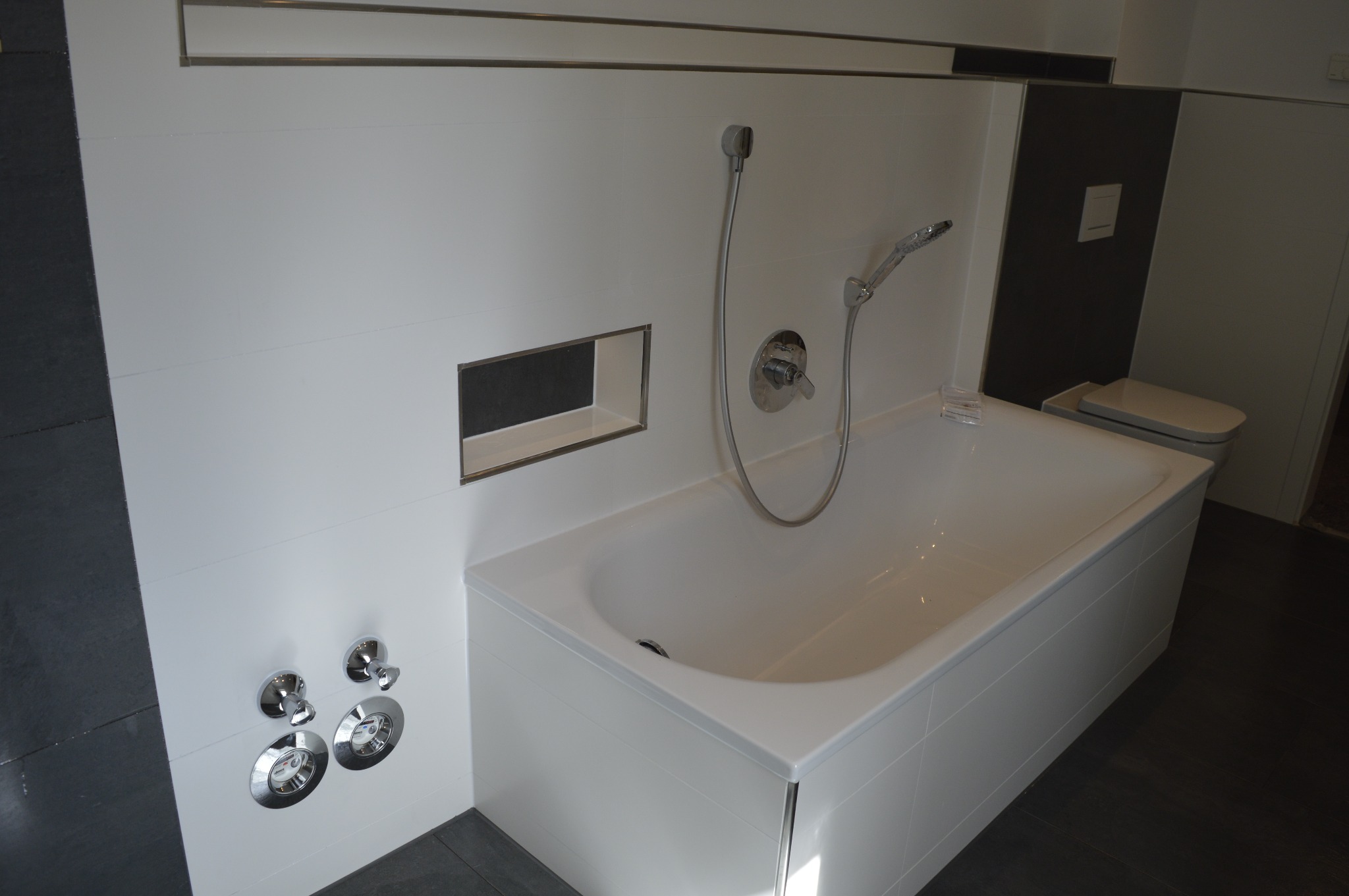 Nowoczesna biała wanna z chromowaną armaturą i wnęką w ścianie wykończonej płytkami, widok z góry na wyremontowaną łazienkę z ciemną podłogą i częściowo widoczną toaletą.