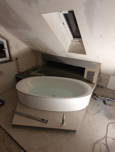 Owalna wanna wolnostojąca, częściowo napełniona wodą, w trakcie montażu w łazience na poddaszu ze skośnym oknem dachowym; widoczne narzędzia i materiały budowlane.