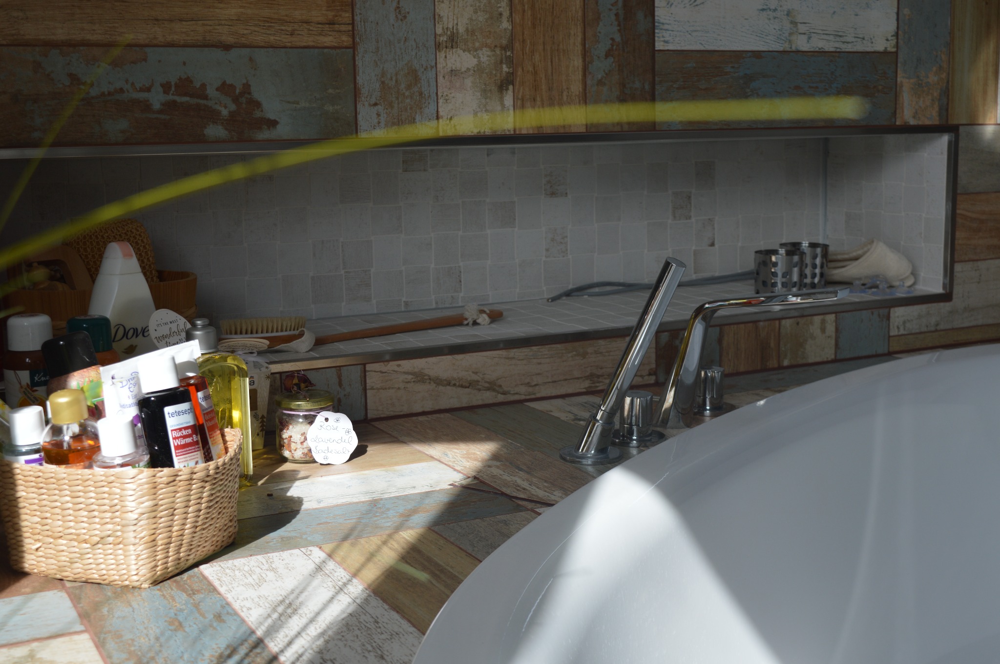 Fragment łazienki z wanną, baterią i półką wnękową wyłożoną mozaiką, na blacie wiklinowy koszyk z kosmetykami i słoik z etykietą Rose-Lavendel.