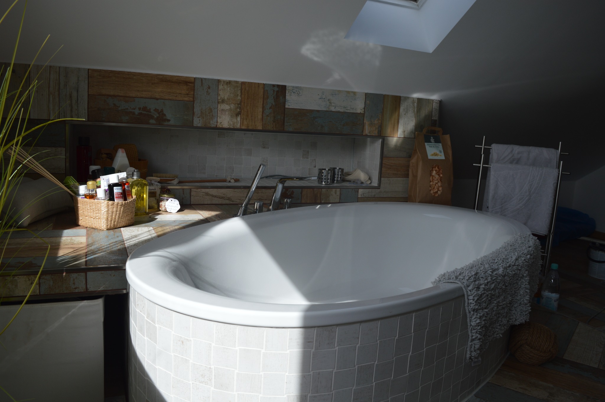 Owalna wanna obłożona mozaiką w jasnej łazience na poddaszu z oknem dachowym, dekoracyjna ściana z drewnopodobnych paneli, wiklinowy koszyk z kosmetykami i brązowa papierowa torba w tle.