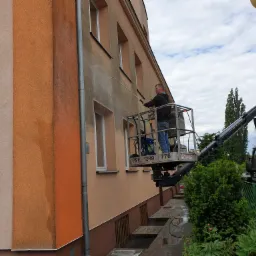 Pracownik na podnośniku koszowym spryskuje wodą elewację budynku, widoczne różnice w kolorze elewacji przed i po czyszczeniu.