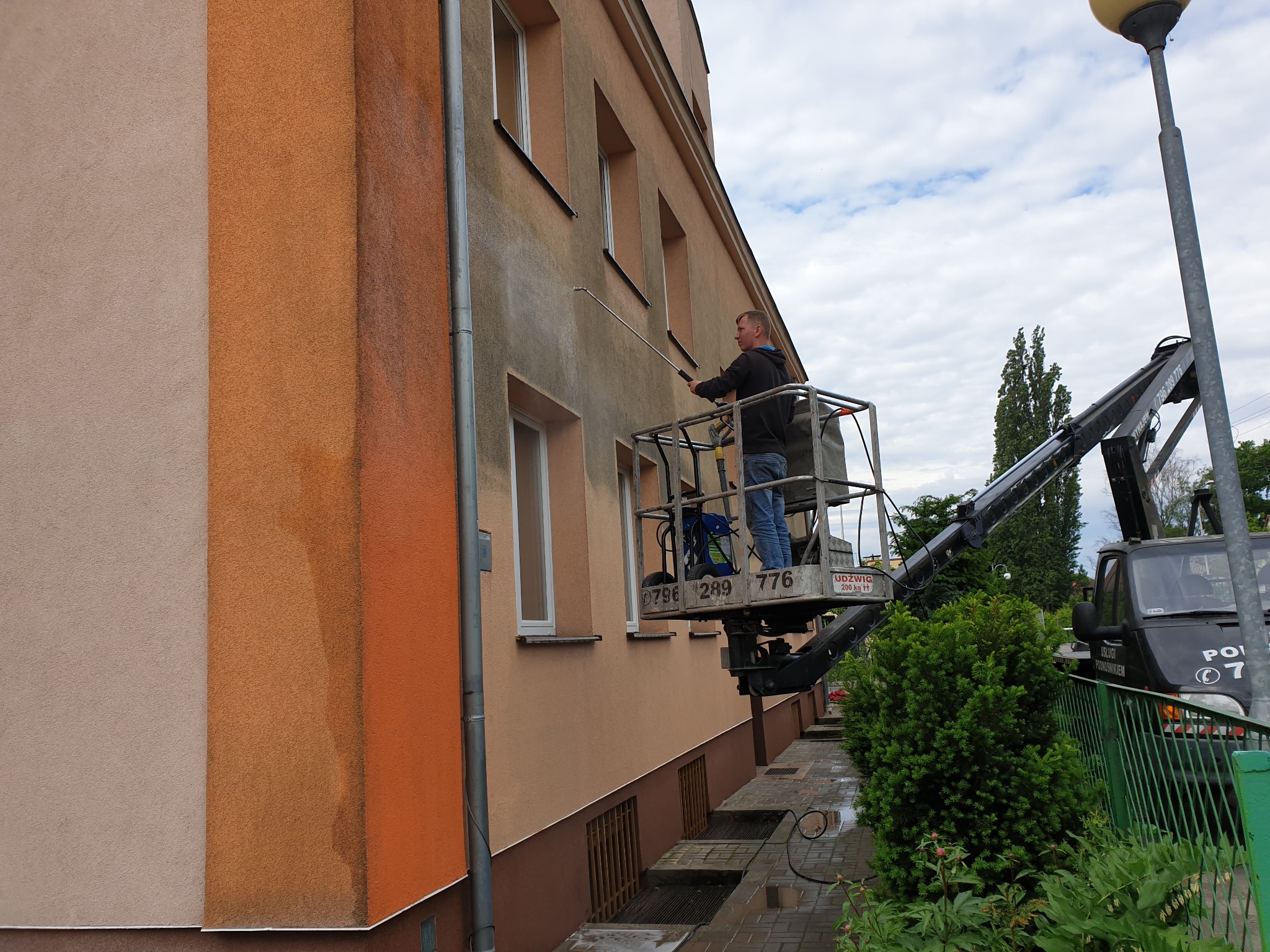 Pracownik na podnośniku koszowym spryskuje wodą elewację budynku, widoczne różnice w kolorze elewacji przed i po czyszczeniu.