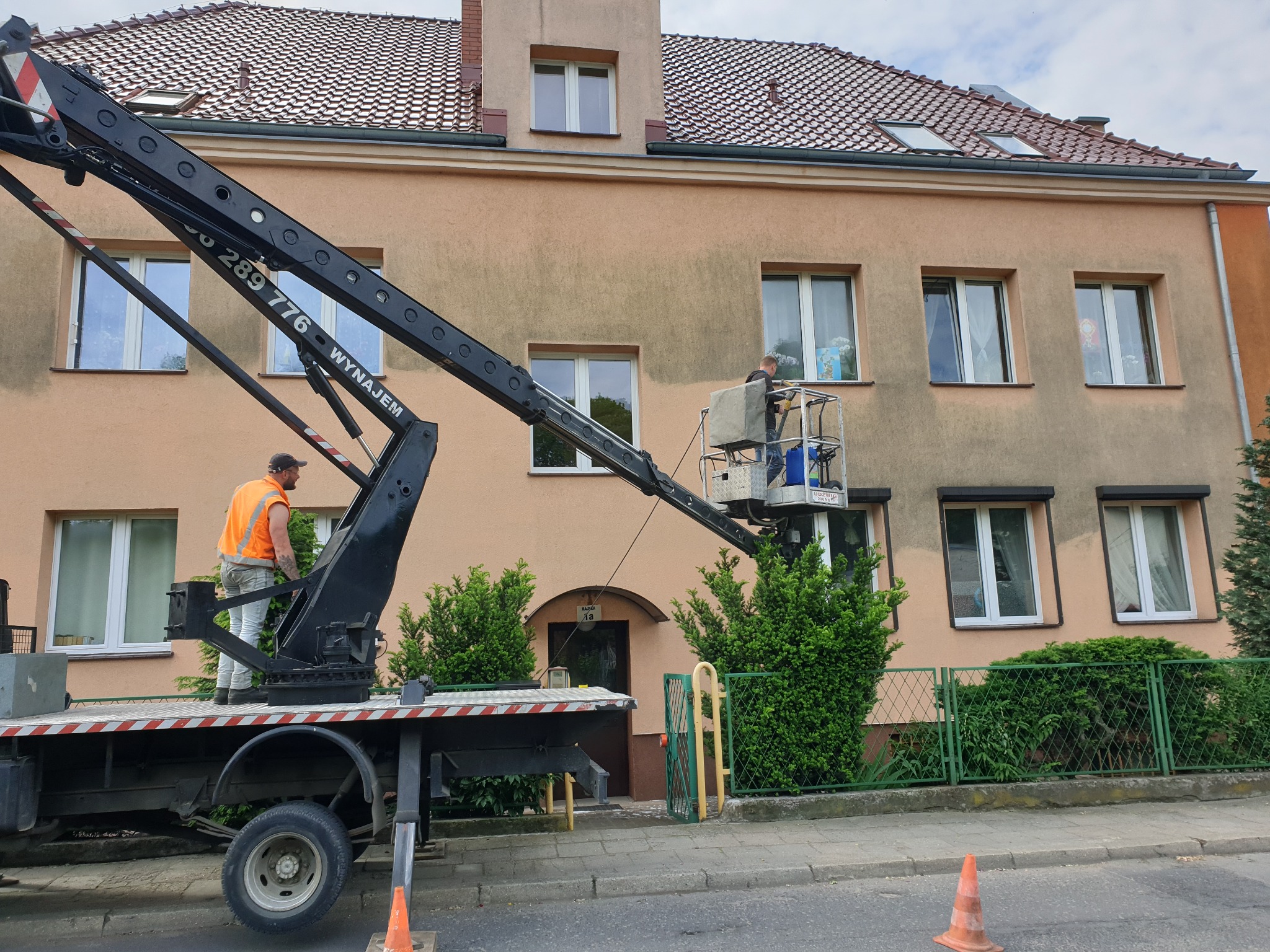 Pracownik w pomarańczowej kamizelce, stojący na platformie podnośnika koszowego, czyści okna na wysokości w budynku o beżowej elewacji, widoczne krzewy i ogrodzenie.