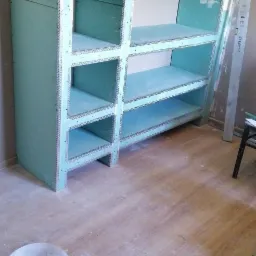 Konstrukcja regału z płyty karton gips z metalowymi narożnikami, widoczne narzędzia na górnej półce, wiadro i skrzynka z narzędziami na podłodze.