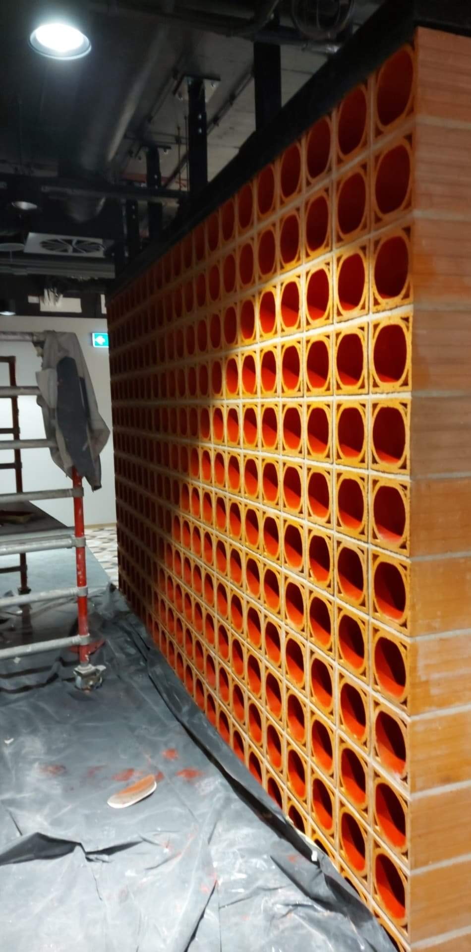 Fragment ściany z dekoracyjnych, pomarańczowo-beżowych pustaków ceramicznych w kształcie kwadratów z okrągłymi otworami, zabezpieczonej folią malarską, widoczny fragment rusztowania.