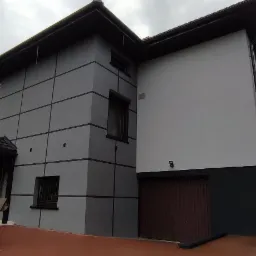 Dwukondygnacyjny dom po remoncie elewacji, z szarą fasadą podzieloną na kwadraty i białą ścianą, widoczny podjazd i częściowo samochód dostawczy.