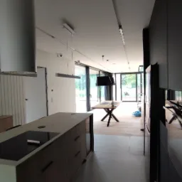 Nowoczesna kuchnia z wyspą i dużymi oknami, widok na zewnątrz, minimalistyczny design, czarne szafki i okap, drewniany stół z czarnymi nogami w tle.