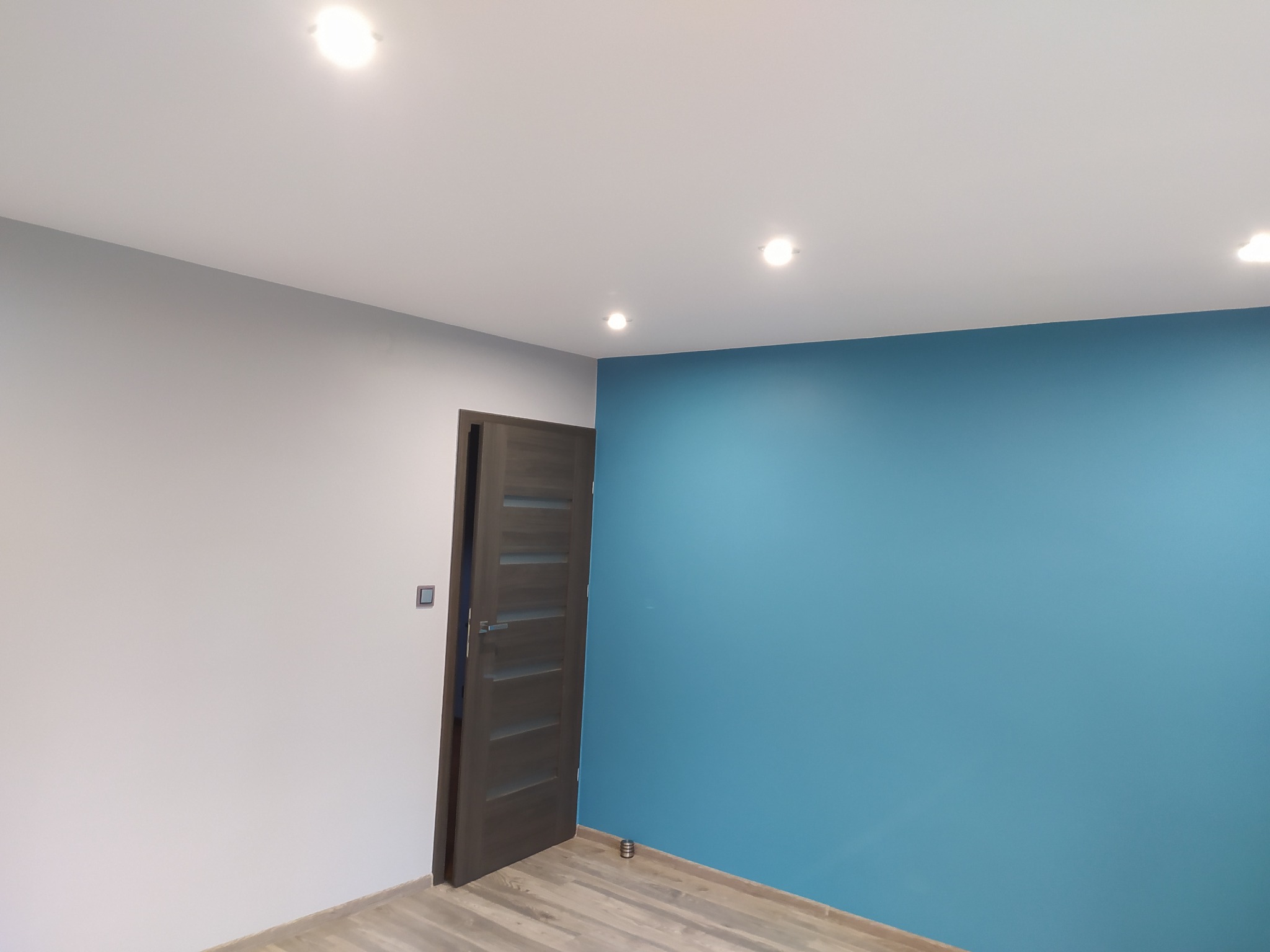 Pokój po malowaniu ścian na szaro i turkusowo, z drewnianą podłogą i ciemną, przeszkloną, otwartą ościeżnicą drzwiową.