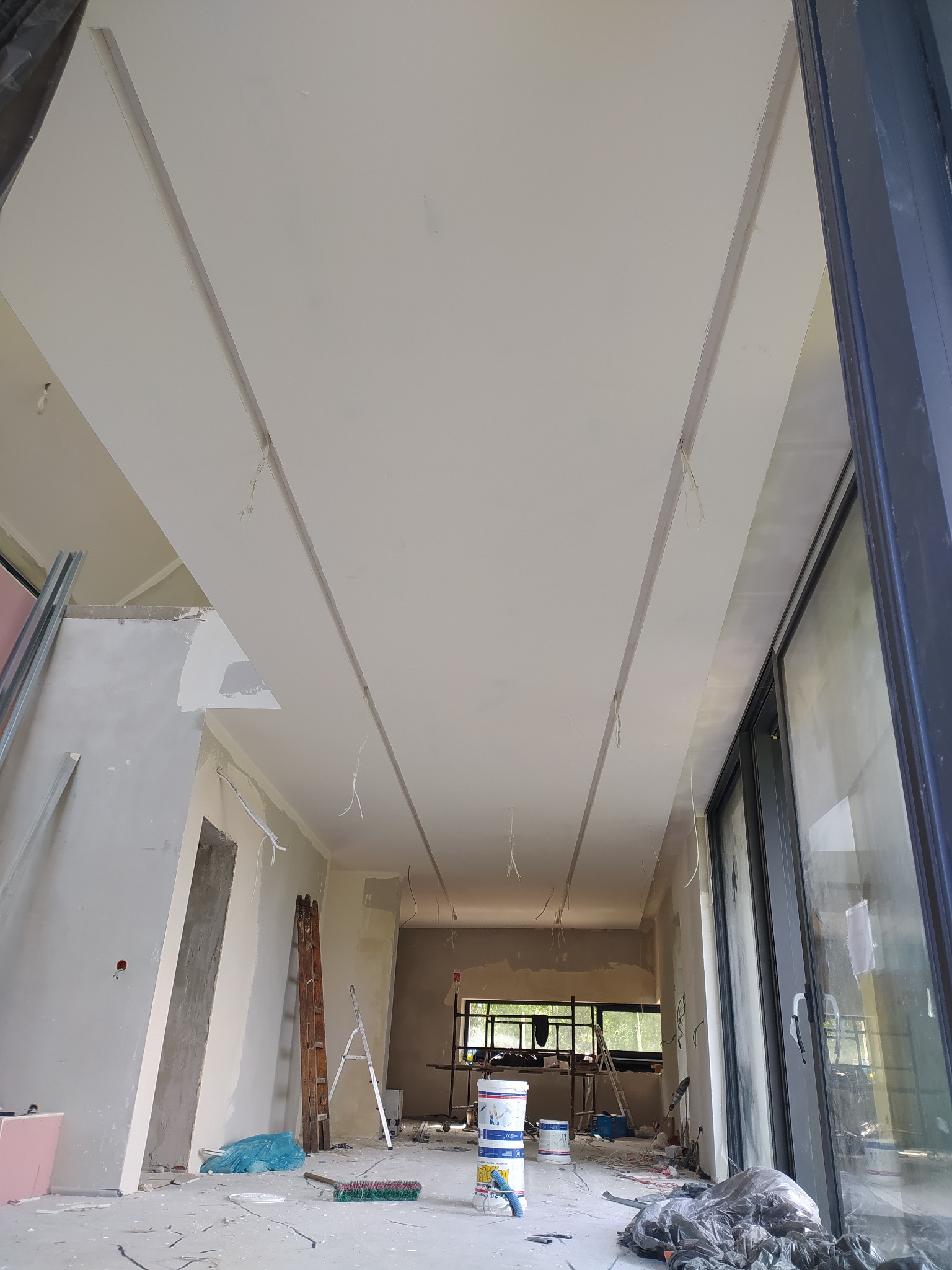 Wnętrze w trakcie prac wykończeniowych: sufit z zamontowanymi profilami, ściany częściowo otynkowane, widoczne drabiny, puszki z farbą i narzędzia na podłodze.