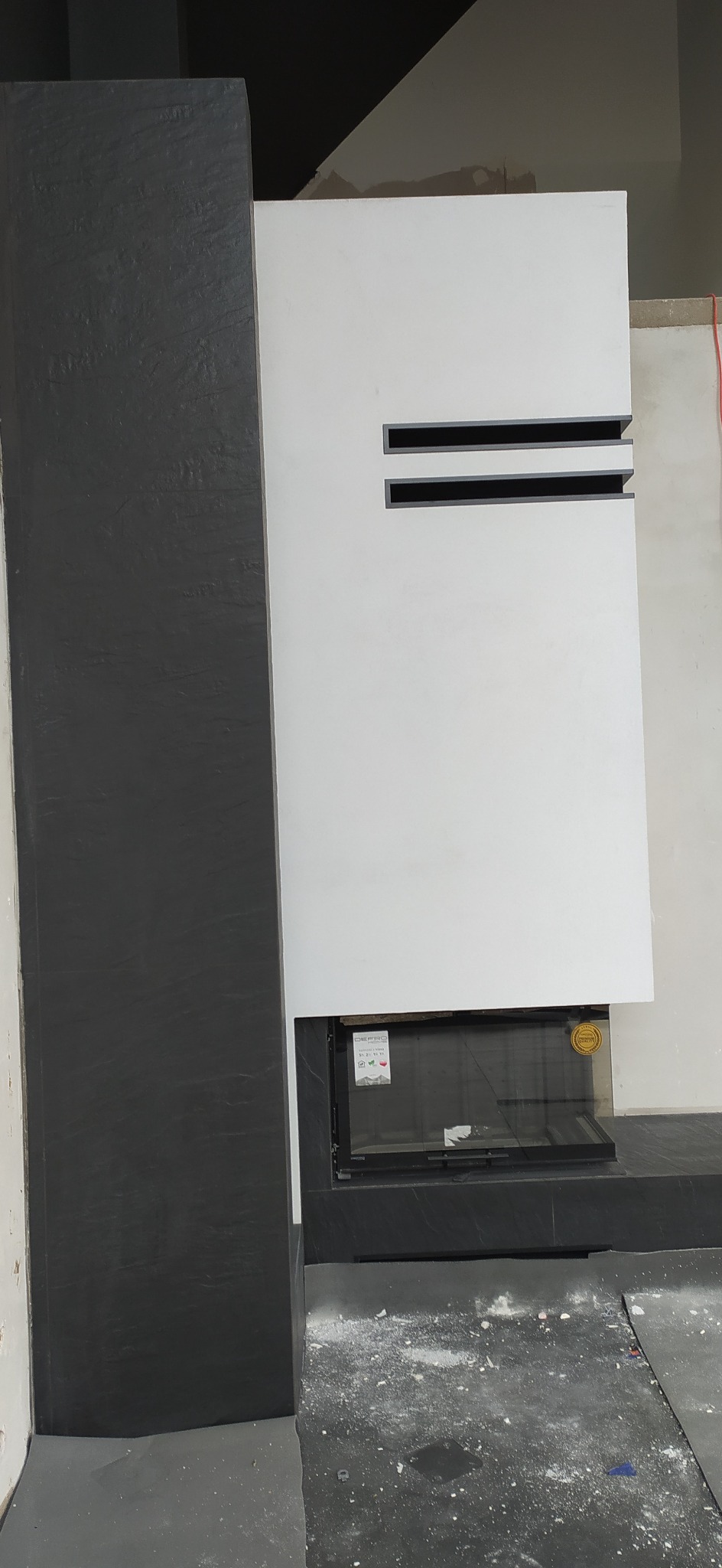 Nowoczesny kominek obudowany płytami w kolorze białym i grafitowym, z widocznymi szczelinami wentylacyjnymi i szybą kominkową, w trakcie prac wykończeniowych.